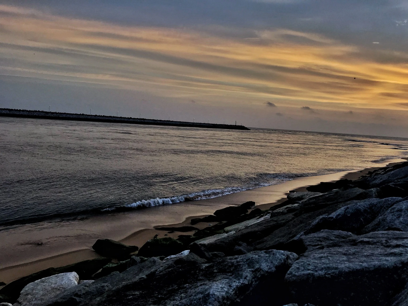 Sunset over the sea at Gangolli with a rocky breakwater silhouette and a band of orange cloud reflected on wet sand