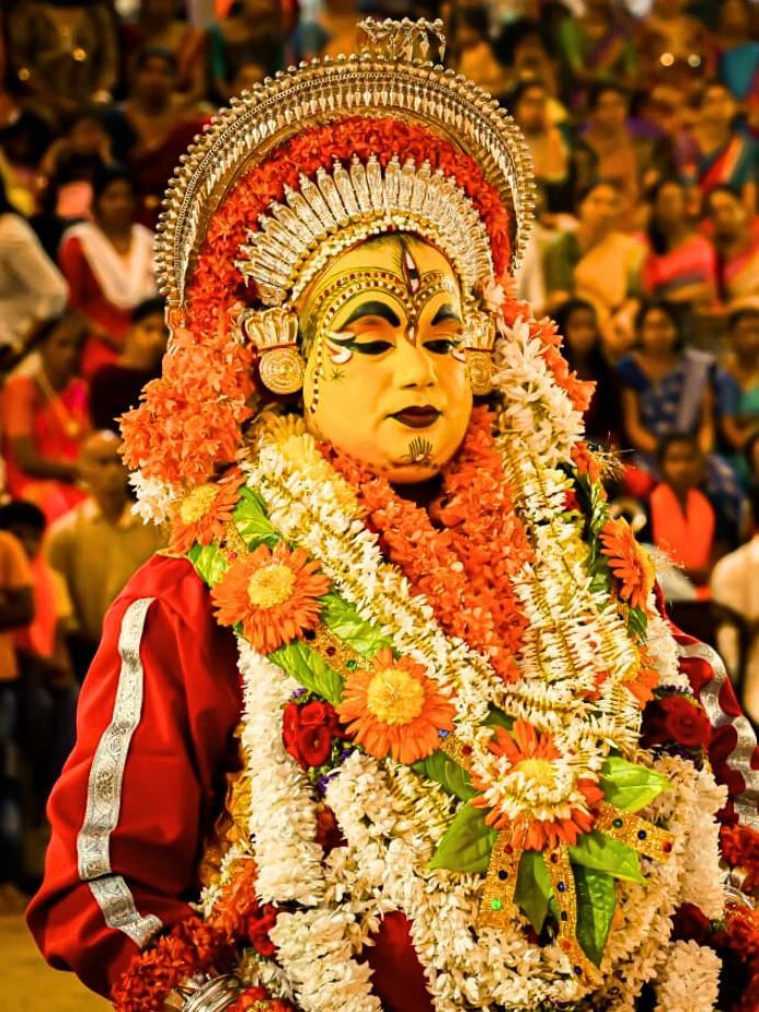 A Bhoota Kola performer wearing a tall silver crown, painted yellow face and a vast garland of orange and white flowers, standing amid a crowd of devotees