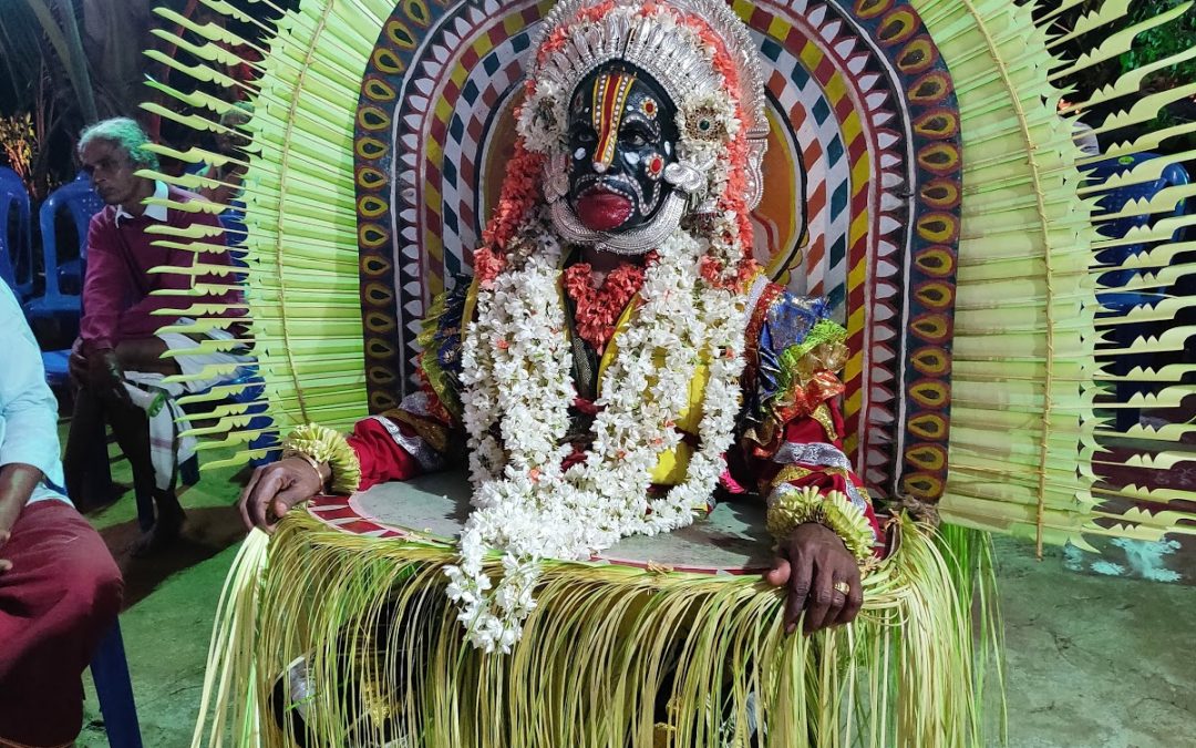 A Bhoota Kola performer with a painted face seated before a fan of tender coconut fronds, draped in flower garlands